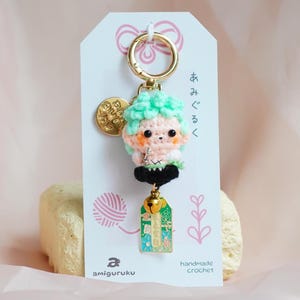 May include: A green and pink crocheted keychain with a bell and a tag that says "handmade crochet" and "amigurumi". The keychain is attached to a gold key ring.