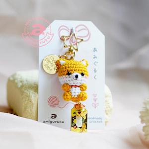 May include: A yellow crocheted amigurumi fox keychain with a white chest and red collar. The keychain has a gold star charm, a gold charm with Japanese text, and a gold charm with a bell and a small gold fish.