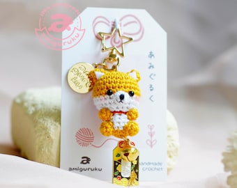 May include: A yellow crocheted amigurumi fox keychain with a white chest and red collar. The keychain has a gold star charm, a gold charm with Japanese text, and a gold charm with a bell and a small gold fish.