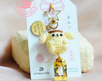May include: A handmade amigurumi keychain featuring a yellow crocheted character with a brown hat and black eyes. The keychain includes a gold star clasp, a round gold charm, and a decorative tag with Japanese text and a bell charm. The amiguruku logo is visible.