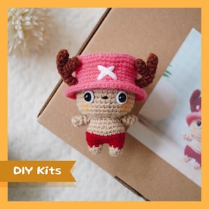 May include: A crocheted plush toy of a cartoon character with brown antlers, a pink hat with a white X, and red shorts. The toy is sitting on a brown box with the text "DIY Kits" on a white ribbon.