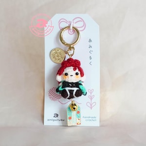May include: A handmade crochet keychain featuring a small character with red hair, black and green clothing, and a gold-colored bell charm. The keychain includes a gold ring and a small tag with Japanese text and the word "handmade crochet".
