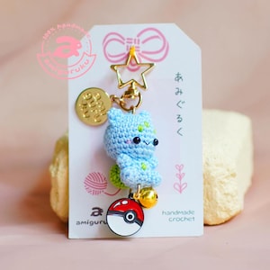Op de afbeelding: Een handgemaakte gehaakte sleutelhanger met een lichtblauw amigurumi-personage met groene accenten, een gouden ster en een Pokeball-bedel. Het label luidt "handmade crochet" en "amiguru".