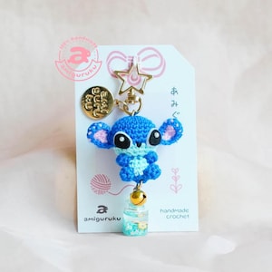 May include: A handmade blue crochet Stitch keychain with a gold star clasp and a small bell. The keychain includes a small bottle filled with blue and white glitter. The tag reads "amiguruku" and "handmade crochet".