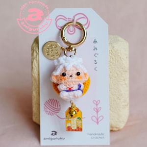 May include: A handmade crochet keychain featuring a white-haired character with a peach face, orange ears, and a blue and red cross design. The keychain includes a gold ring, a small gold charm, and a yellow tag with Japanese text. The tag reads "handmade crochet".
