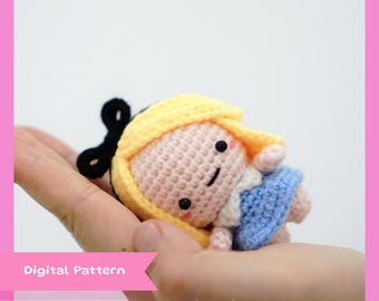 May include: A crocheted doll with blonde hair, a pink dress, and a blue dress. The doll has a black bow on its head and a smiling face. The doll is being held in a hand.