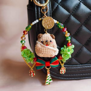 May include: A crocheted pug dog charm with a green and red bow, hanging from a gold chain with a beaded wreath and a gold charm that says "ami guru ku".