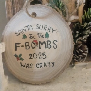 May include: A round, wooden Christmas ornament with the text "SANTA SORRY For the F-BOMBS 2025 WAS CRAZY" in black ink. The ornament has a twine loop for hanging and is decorated with small green trees, a Santa hat, and holly.