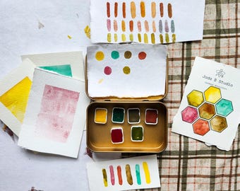 May include: A watercolor paint set with a gold metal case. The set includes 9 watercolor pans, a honeycomb design with watercolor swatches, and several sheets of paper with watercolor swatches in various colors.