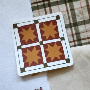 Op de afbeelding: Een vierkante sticker met een witte rand heeft vier stervormige ontwerpen in een rasterpatroon. De steruitbarstingen zijn oranje, tegen een bordeauxrode achtergrond. De sticker ligt op een witte en geruite stof.