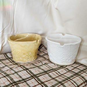 Puede incluir: Dos ceniceros de cerámica. Uno es amarillo pálido con un patrón de panal y floral. El otro es blanco con un patrón de panal. Ambos tienen un interior dividido y una muesca para apoyar objetos. Los ceniceros están sobre una tela a cuadros.