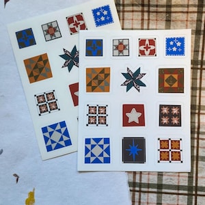 May include: Two sheets of stickers featuring colorful quilt block designs. Each sticker has a square shape with various geometric patterns in shades of blue, orange, brown, and red. The stickers are on a white background, with a plaid fabric in the background.