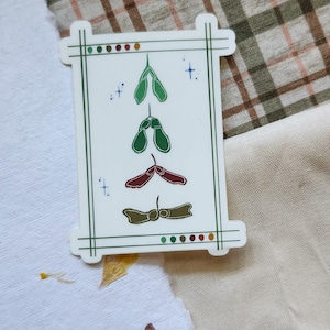 Op de afbeelding: Een witte sticker met een groene rand toont gestileerde zaadillustraties in groen, rood en bruin. De sticker heeft een rechthoekige vorm met een decoratieve rand en kleine stippen.