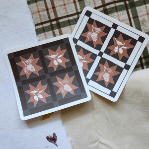 Op de afbeelding: Twee vierkante kaarten met een herhalend patroon van bruine sterren en bloemmotieven op een zwart-grijze geblokte achtergrond. De kaarten hebben witte randen en worden weergegeven op een getextureerd oppervlak.