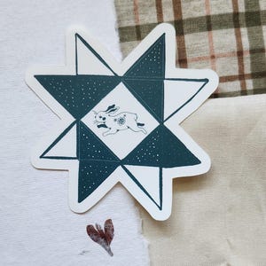 May include: A star-shaped sticker with a geometric design in teal and white. The center square features a running rabbit illustration. The sticker is on a white surface with a plaid fabric in the background. A small, red-brown leaf shape is also visible.