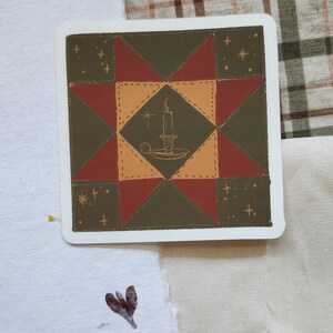 Op de afbeelding: Vierkante sticker met een quiltblokontwerp. De sticker heeft een centrale ruit met een kaarsillustratie, omringd door driehoeken in tinten rood, bruin en olijfgroen. Kleine ster- en kruisdetails zijn verspreid over de randen.