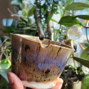 Puede incluir: Un pequeño cuenco de cerámica hecho a mano con un borde ancho y acampanado que presenta una muesca. El esmalte exterior transita de una base blanca a un acabado moteado en tonos bronce y marrón con vetas azul iridiscente y puntos oscuros.