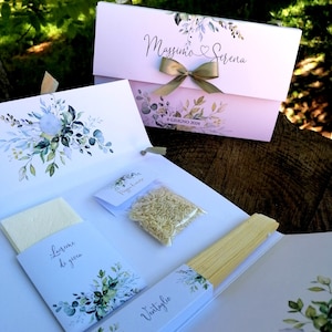 Può includere: Una scatola di bomboniere da matrimonio con sfondo bianco e design floreale verde. La scatola è legata con un nastro dorato e ha i nomi "Massimo" e "Serena" stampati sul davanti. La scatola contiene una piccola busta bianca con le parole "Rice & Love" stampate su di essa, un piccolo sacchetto di riso, una busta bianca con le parole "Tears of Joy" stampate su di essa e un ventaglio bianco con la parola "Ventilaggio" stampata su di esso.