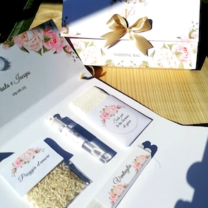 Può includere: Una borsa regalo di nozze bianca con un design floreale e un nastro dorato. La borsa contiene una piccola bottiglia di bolle, un pacchetto di riso, un fazzoletto e un ventaglio. La borsa ha anche un'etichetta che dice "Wedding Bag".