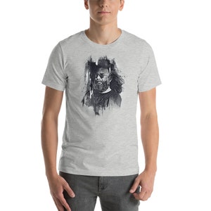 May include: A light gray t-shirt featuring a black and white artistic portrait of a man with a beard and sunglasses. The graphic has a painterly, abstract style. The shirt is short-sleeved and has a crew neck.
