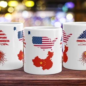 May include: Three white ceramic mugs with a design of the American flag and the Chinese flag. The American flag is depicted as a tree with roots that are the Chinese flag. The mugs are on a wooden surface.