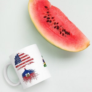 May include: White ceramic mug with a tree design featuring the American and Haitian flags. The tree's roots are in the Haitian flag and the branches are in the American flag.