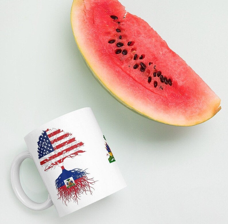 May include: White ceramic mug with a tree design featuring the American and Haitian flags. The tree's roots are in the Haitian flag and the branches are in the American flag.