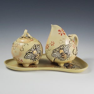 May include: A ceramic cream and sugar set with a tray. The set features a sugar bowl with a lid, a creamer, and a tray, all decorated with bee and honeycomb designs. The pieces are light yellow with red and black accents.