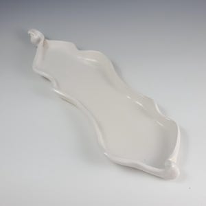 May include: A white, elongated ceramic serving tray with a wavy, organic shape. The tray has a smooth, glossy finish and a shallow depth, perfect for displaying appetizers or small items. The design features a subtle, flowing form.