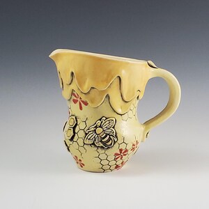 May include: A ceramic pitcher with a yellow glaze, featuring a bee and honeycomb design. The top edge has a dripping honey effect. Red flower accents are scattered across the surface. The handle is curved and attached to the side.