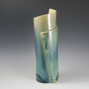 May include: A tall, ceramic vase with a unique design. The vase is primarily blue with green and tan accents. A diagonal section is attached with three circular details. The top curves outward. The vase is approximately 10 inches tall.