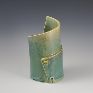 May include: A handcrafted ceramic vase with a unique, curved design. The vase features a blend of green and blue hues with a glossy finish. The design includes a folded section secured with small, circular accents, adding a modern touch to the pottery.
