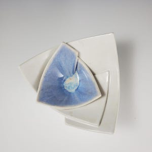May include: A stack of three ceramic dishes in a geometric design. The bottom two are white, square-shaped with rounded corners. The top dish is a blue triangle with a lighter center, creating a layered effect. The dishes are arranged on a white background.