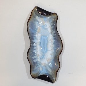 May include: An elongated ceramic tray with a wavy, organic rim. The interior features a reactive glaze with a central band of light blue transitioning into white and dark brown at the edges. The glossy surface displays a mottled, fluid pattern.