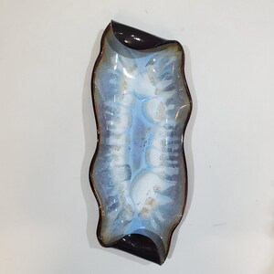 May include: An elongated ceramic tray with a wavy, organic rim. The interior features a blended glaze of white, light blue, and dark blue, with subtle golden-brown speckles. The outer edges and ends are finished in a solid, glossy dark brown glaze.