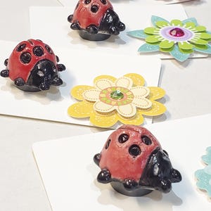 May include: A collection of handmade ladybug and flower-shaped decorative magnets. The ladybugs are red with black spots and heads. The flowers are pink, yellow, and green with gem centers. They are arranged on white cards.