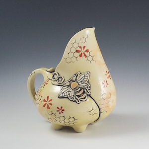 May include: Cream-colored ceramic pitcher with a bee and honeycomb design. The pitcher features a bee applique, red flower accents, and a honeycomb pattern. It has a handle and three small feet. The pitcher is approximately 7 inches tall.