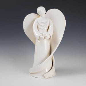 May include: A white ceramic angel figurine with outstretched wings and a flowing gown. The angel holds a small flower in its hands. The sculpture has a smooth, glossy finish and is set against a gradient background.