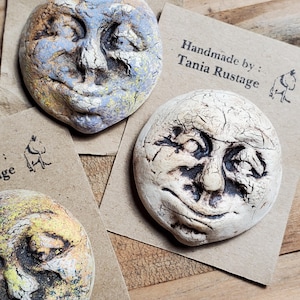 May include: Handmade ceramic moon face sculptures in various colors. The sculptures have a textured surface and are in shades of blue, yellow, and cream. Each sculpture has a unique facial expression. The artist's name, Tania Rustage, is printed on a brown card.