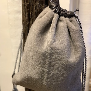 Könnte beinhalten: Eine graue Kordeltasche aus weichem, flauschigem Stoff mit einem schwarzen Kordelzug und schwarzen Kordelgriffen.