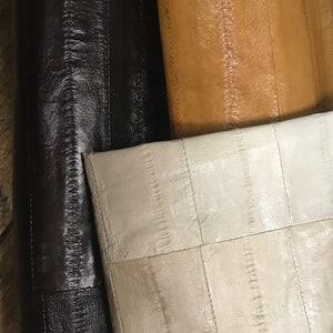Op de afbeelding: Close-up van drie rollen leer in verschillende kleuren. De eerste rol is donkerbruin, de tweede is bruin en de derde is gebroken wit. Elke rol is gestikt met verticale naden.