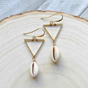 Peut inclure: Une paire de boucles d'oreilles triangulaires dorées avec un pendentif en coquillage cauri. Les boucles d'oreilles ont une fermeture à crochet et sont présentées sur une surface en bois clair. Les coquillages sont blancs avec un intérieur marron.