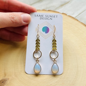 May include: Gold-colored earrings with a teardrop-shaped, opalescent stone. The earrings have a geometric design with arrow-shaped accents and a circular element. The card behind the earrings reads "SAME SUNSET DESIGN".