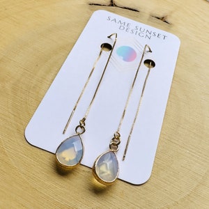 Peut inclure: Boucles d'oreilles pendantes dorées avec des pierres opalescentes en forme de poire. Les boucles d'oreilles sont présentées sur une carte blanche avec le texte "SAME SUNSET DESIGN" et un motif géométrique coloré.