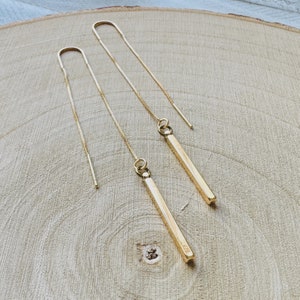 May include: A pair of gold-colored threader earrings. Each earring features a delicate chain that threads through the ear, with a small, rectangular bar pendant hanging at the end. The earrings are displayed on a light-colored wooden surface.