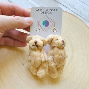 May include: A pair of beige teddy bear earrings. The earrings are made of soft, plush material and feature small brown embroidered details. The earrings are attached to gold-colored hooks and are displayed on a white card with the text "SAME SUNSET DESIGN".