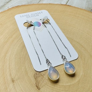 May include: Silver threader earrings with teardrop-shaped, opalescent stones. The earrings are displayed on a white card with the text "SAME SUNSET DESIGN". The stones have a pearlescent glow.