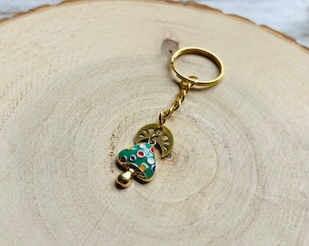 May include: A gold-coloured keychain featuring a green mushroom with white and red polka dots. The mushroom is attached to a gold-coloured crescent moon charm and a gold-coloured key ring.