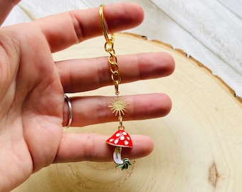 May include: A gold-coloured keyring featuring a red mushroom charm with white spots, a gold sunburst charm, and a gold chain. The mushroom has a white stem and green leaves. The keyring is held up by a hand.