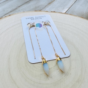 May include: Gold-tone chain earrings with white and blue teardrop-shaped pendants. The earrings are displayed on a white card with the text "SAME SUNSET DESIGN". The background is a light wood grain.